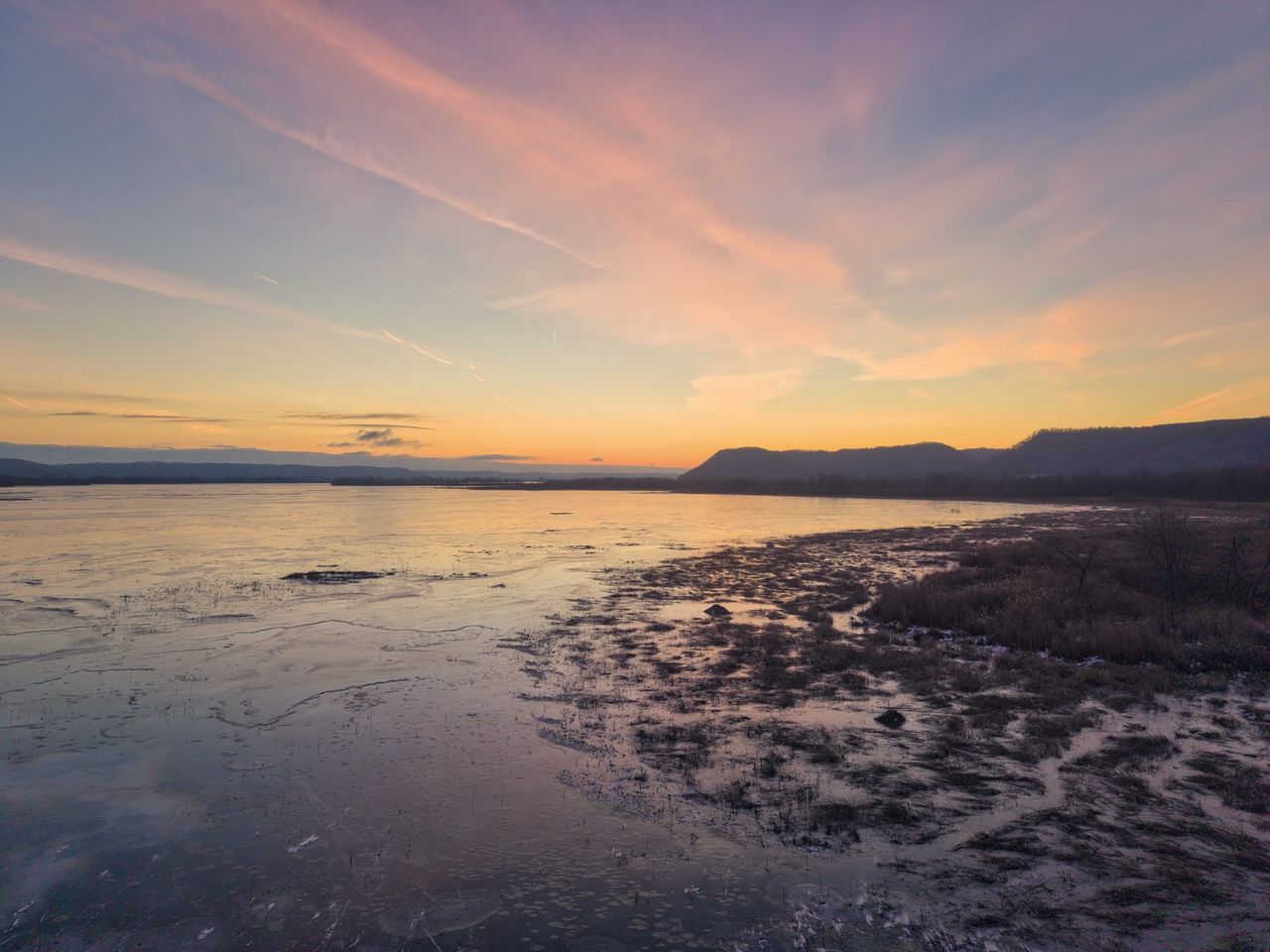 Fleuve gelé au crépuscule, ciel rose et orangé au-dessus d'une rive de marais